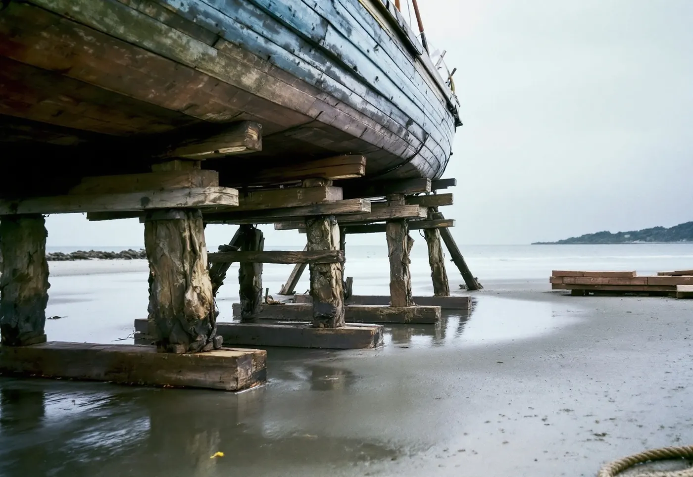 Timber blocking and angled shoring supporting a wooden hull on a wet sand foreshore as the tide returns